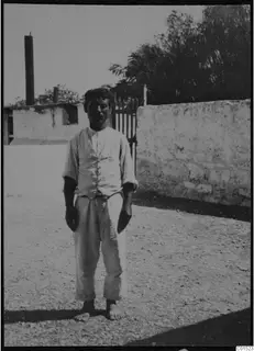 Man in long trousers, waistcoat and bare feet standing in a yard in front of a whitewashed wall