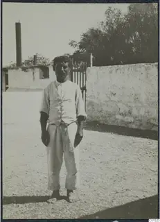 Man in shirtsleeves, waistcoat and long trousers standing barefoot on a gravel yard in front of a whitewashed wall.