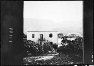 View through a window of a white two-storey house with a balcony, orchard trees in front and mountains behind.