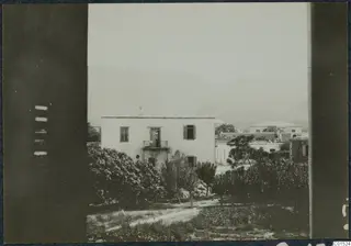 View through a window frame over a whitewashed two-storey house, trees and mountains behind it