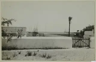 White plastered wall and wooden gate in the foreground, with masted boats and the sea beyond, at Larnaca