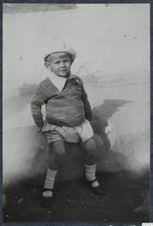 Young child in a knitted sweater and pale cap sitting on a large stone against a whitewashed wall.