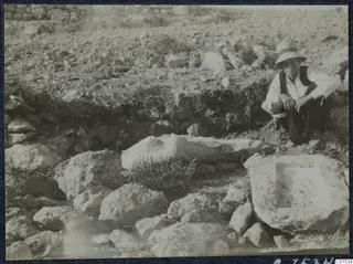 Carved stone fragment and a block with a recess among rubble at Vouni, with a seated man beside them.