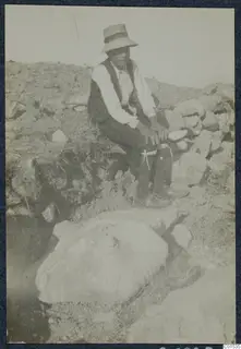 Person in a wide-brimmed hat seated beside a large stone block in a trench at Vouni