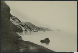 Rocky coastline with a cliff, a shingle shore and calm sea, looking out toward distant headlands.