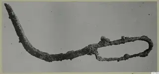 Corroded metal object from Vouni: a long curved arm tapering to a point, ending in a slender oval loop with small knobs.