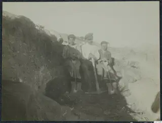 Solon and two workers sitting in an excavation trench, holding spades and a pick