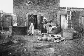 Packing pottery in a mud-brick courtyard at Milia, with a man by a doorway and a woman carrying a bundle.