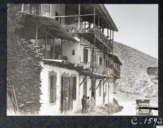Whitewashed house with timber balconies and a stacked wood wall in Kalopanayotis, with people standing at the doorway