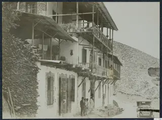 Two-storey house with timber balconies and shuttered windows in Kalopanayotis village, with people standing at the doorway.