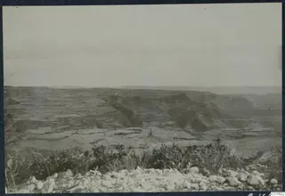 Wide view over terraced hillsides and a deep valley, with scrub and stones in the foreground under a pale sky.