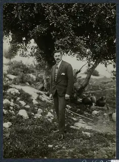 Lazaros in a dark suit standing under a large tree at Vouni, with jars on the ground to the right.