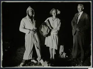 Three people posing outdoors at night: a man in a striped robe, a woman carrying blankets, and a man in a suit and tie.