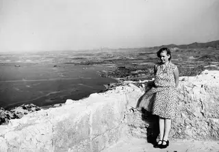 Miss Atterman in a spotted dress standing on a stone wall at Vouni, overlooking the sea and the plain.