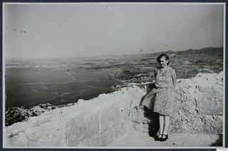 Miss Atterman in a patterned dress standing on a stone wall at Vouni, with the sea and coastal plain behind her.