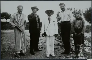 Five people standing in a row on a gravel track at Vouni, one in a striped hooded robe and one in a white suit.