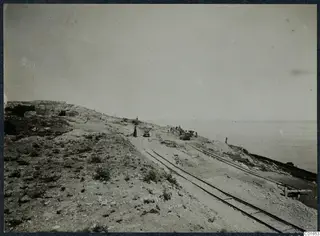 Narrow-gauge rails and small wagons running across a rocky slope above the sea, with cut stone walls at the left and several people.