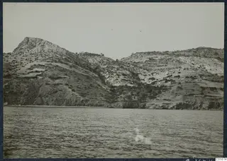 View from the sea of a rocky, scrubby headland rising above a cliff-lined shore at Vouni