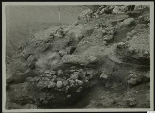 Excavation at Petra tou Limniti with exposed stones and a vertical ranging pole on a sloping earth surface