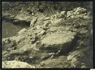Excavation among rocks at Petra tou Limniti, with water to the left and a thin wooden pole at the edge