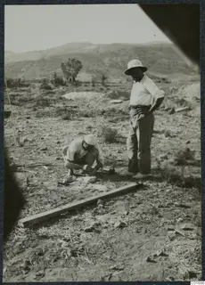 Two men at work on a dry hillside, one crouching and one standing beside a wooden plank on the ground.