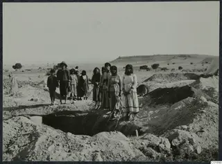 Group of women, a man and a child standing beside an excavated dromos at Ayios Iakovos, with spoil heaps around.
