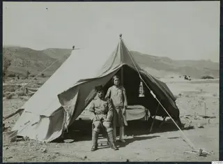 Policeman seated outside a large canvas tent at Ayios Iakovos, with a young girl standing beside him.