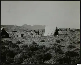 People and white canvas tents on open scrubland at Ayios Iakovos, with mountains behind