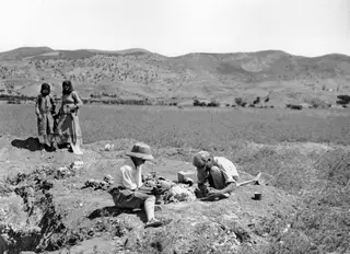 Two men work among skulls on a raised mound at Ayios Iakovos, with two women in long dresses standing behind them