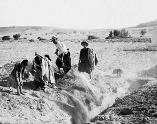 Workers filling in a cut trench at Ayios Iakovos with soil, one man holding a hoe, a tent at the right edge.