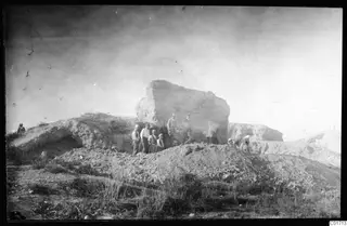 Men stand in front of a large rock face at Bamboula, Kition, during its demolition.