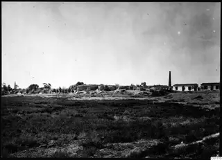 Bamboula from the north: a low stone mound across an open grassy field, with buildings and a chimney to the right