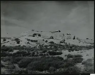 Bamboula mound seen from the southeast, with several people sitting on its top edge above low scrub.