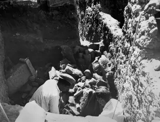 Two men digging among broken stone sculptures in a deep excavation trench at Kition