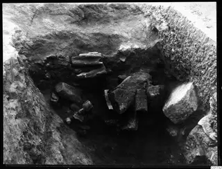 Excavated pit with broken stone pieces lying in situ in dark soil, and a large pale block at the right edge