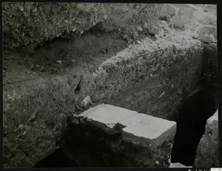 Deep excavation trench with a tall soil section and a large pale stone block at its base, Kition
