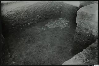 Excavated trench corner at Kition, looking down into a pit with a pale remnant of floor surface on the bottom.