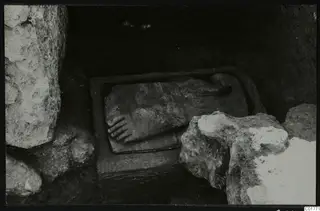 Carved stone figure lying in a rectangular stone trough, with large rough stone blocks around it, at Kition