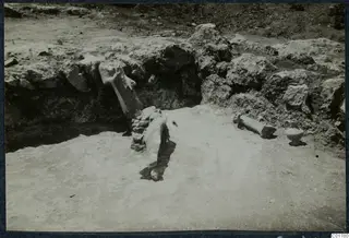 Finds in situ during excavation at Vouni: a small pot and carved stone fragments on a sandy floor below a rubble wall.