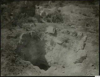 Excavation pit with loose stones and two people among bushes at Kountoura Trachonia
