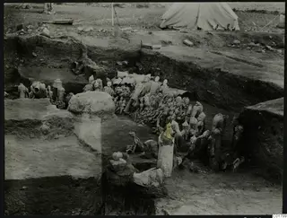 In situ finds in an excavation trench at Ayia Irini, with figurines and vessels and a tent beyond.