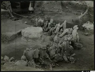 Excavation at Ayia Irini with many figures and vessels lying in the trench, two people standing at the edge