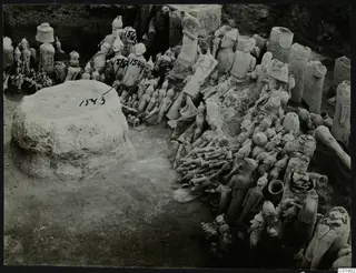 Excavation at Ayia Irini showing a large rounded block and many figures and sculptures heaped among the soil.