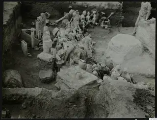 In situ figurines and vessels lying in an excavation trench at Ayia Irini, with a person standing at the edge