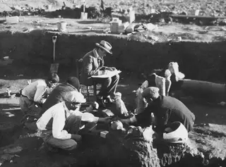 Excavation at Ayia Irini: workers clean sculpture fragments while a seated man sketches beside them in a trench.