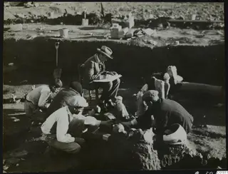 Excavation at Ayia Irini: a seated man in a hat writes while four workers clean and sort broken finds on the ground