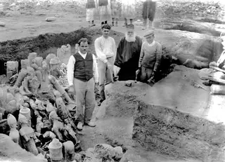Four men stand at the edge of an excavation at Ayia Irini, with terracotta figurines and vessels heaped in the trench to the left.