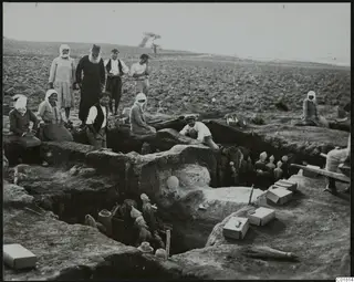 Excavation trench at Ayia Irini with figurines and jars lying in the cut, watched by a group of people at its edge.