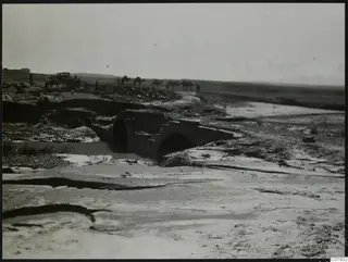 Stone arched bridge over a muddy watercourse at Chiloura, with a line of camels and people on the distant ground.