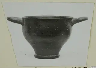Black glazed skyphos with two horizontal handles, photographed against a pale background on a card mount